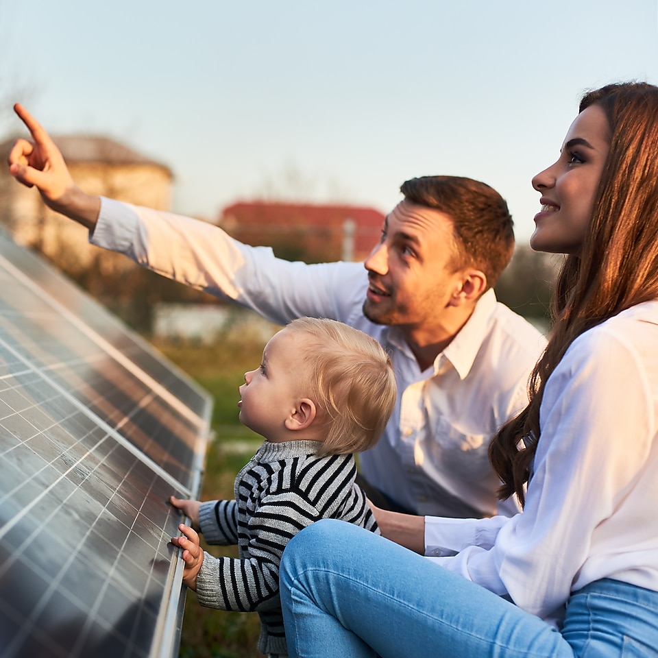 Frau, Mann und Kind knien vor Solarplatten. Mann zeigt mit einem Finger nach oben.