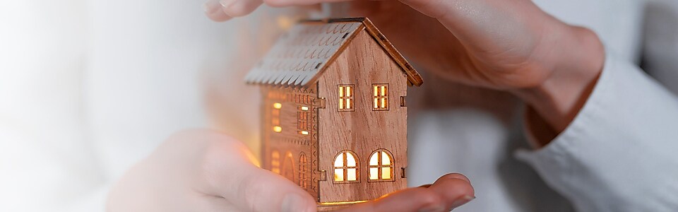 Person hält ein beleuchtetes Miniaturholzhaus schützend in den Händen